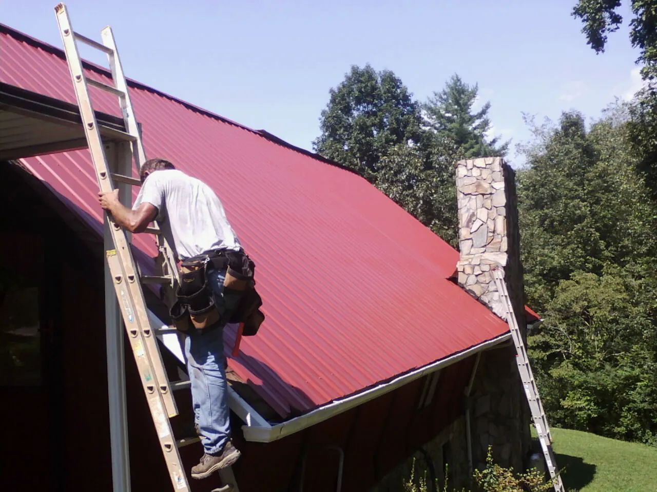 Roofer on a ladder beside a red metal roof and stone chimney — Soffit & Fascia Repair by Roofing Brothers in Honesdale